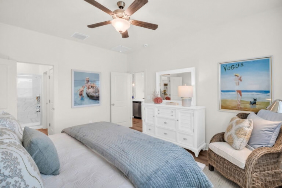 Staged owner's bedroom in a light, airy coastal style, featuring a bed with blue and white bedding, a white dresser, a wicker accent chair, a ceiling fan, beach-themed wall art, and a doorway leading to an attached bathroom.