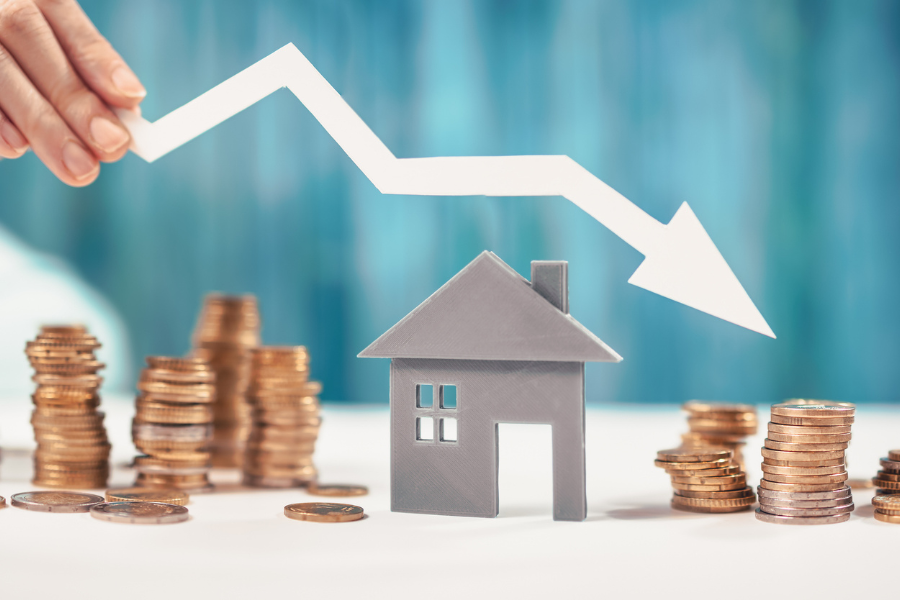 A small gray model house surrounded by stacks of gold coins, with a hand holding a white downward-pointing arrow above the roof, a concept image for declining home prices, mortgage rates, or housing costs.